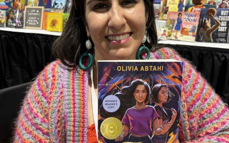 Smiling woman with straight dark hair is holding up the book Twin Flames at the Lee & Low booth at the ALA conference.